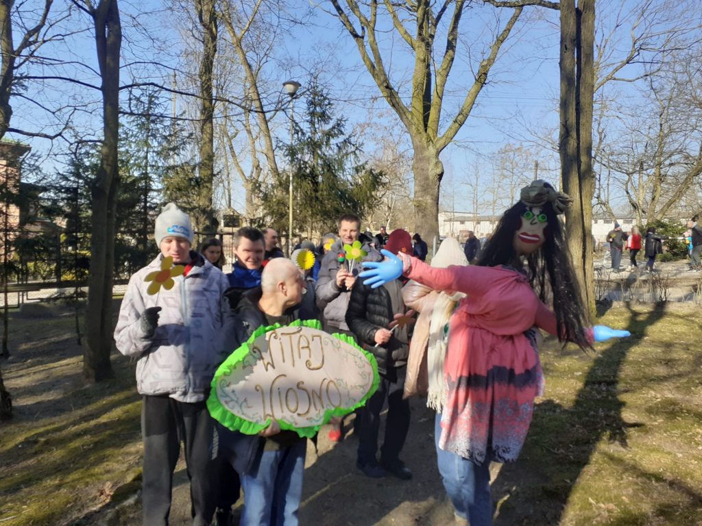 Grupa osób idąca wspólnie, z przodu mężczyzna, który niesie napis Witaj wiosno obok niego osoba trzymająca marzannę