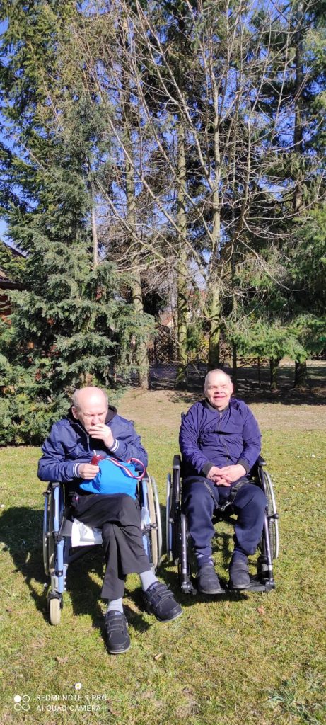 Dwóch podopiecznych siedzi na wózkach inwalidzkich na zielonej trwa za nimi zielone drzewa iglaste. Świeci słońce.