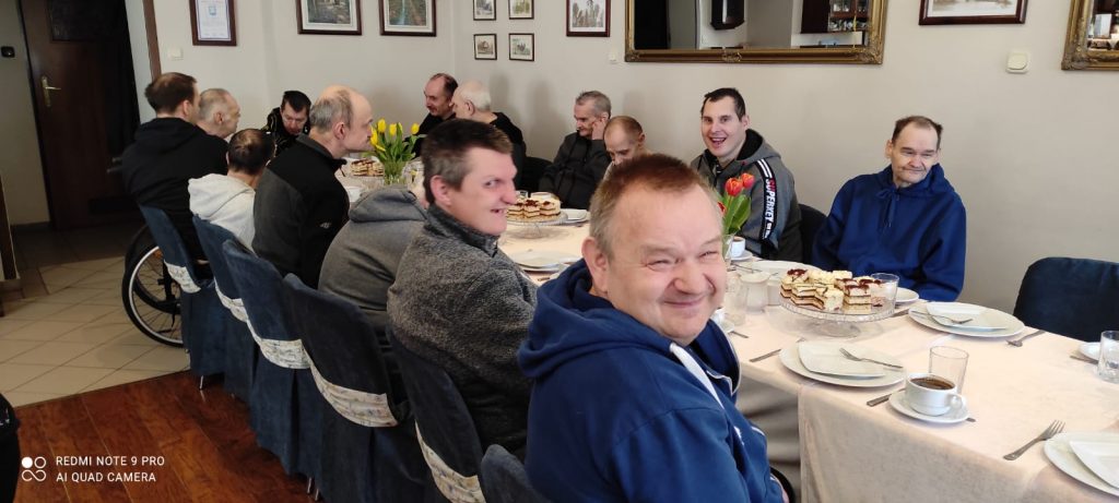 Mężczyzni siedzącą przy stole, trzech z nich ma uśmiech na twarzy. Na stole ciasto, kawa i zastawa do jedzenia