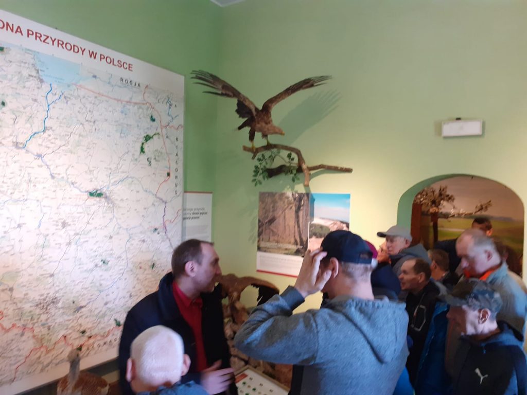 Grupa wycieczkowa wraz z przewodnikiem stoi przed mapą przyrody w Polsce.