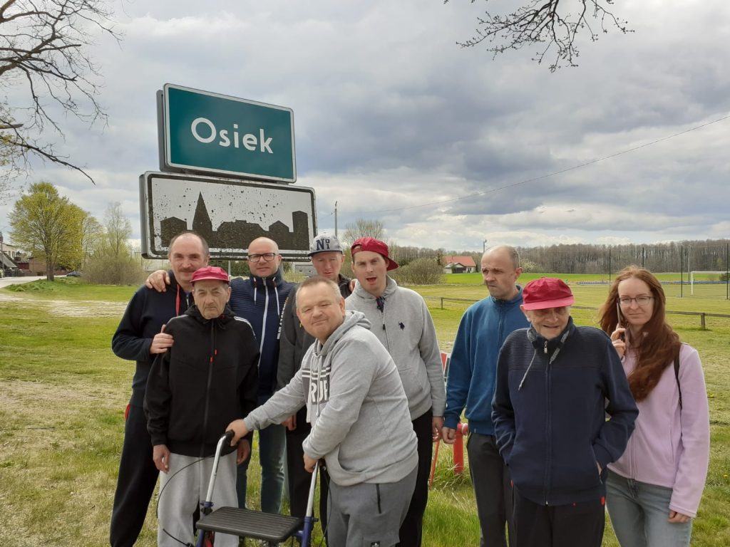Grupa podopiecznych oraz opiekunów stoi pod tablicą na wjeździe do miasta z napisem Osiek. W tle drzewa, łąka i drzewa