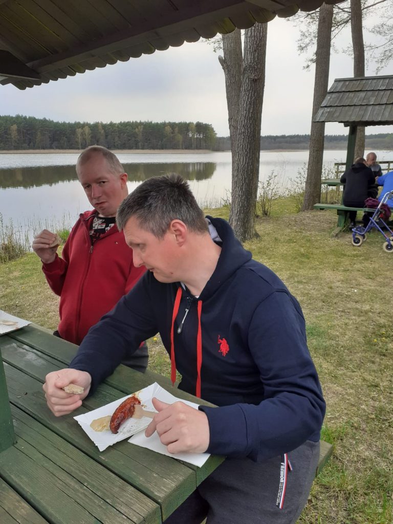 Dwóch mężczyzn siedzi przy drewnianym stole, jedzą kiełbaskę z grilla, za nimi jezioro, a w tafli wody odbija się las.