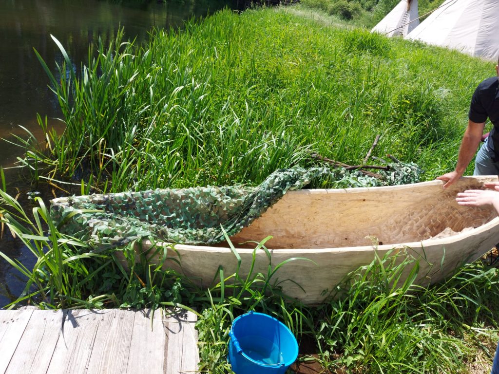 Deska Sup pokryta zielonym kamuflarzem stoi na trawie przy rzece oraz pomoście..