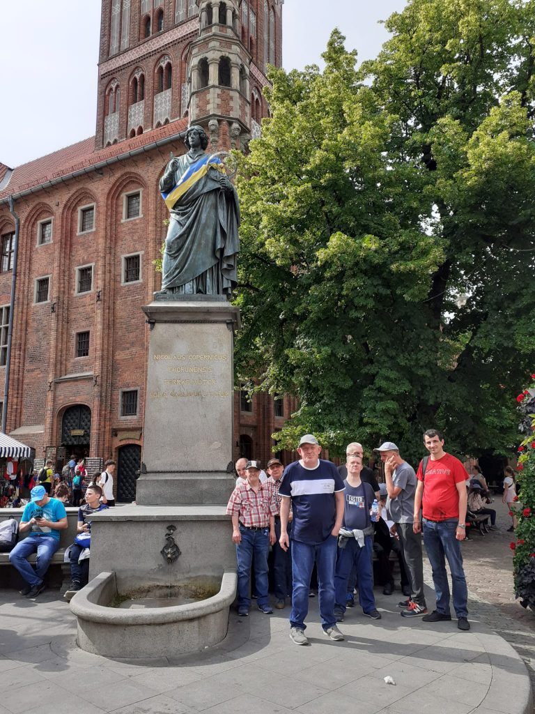 Grupa mieszkańców stojąca przy pomniku Mikołaja Kopernika. Pomnik umieszczony na kolumnie. W tle kościół.