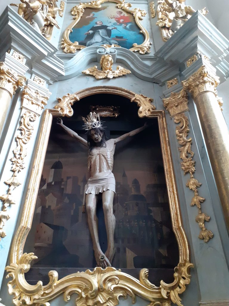 Rzeźba Jezusa konającego na krzyżu, z koroną cierniową na głowie. W koło złote rzeźbienia na ścianie. Nad Jezusem obraz w pozłacanej ramie.