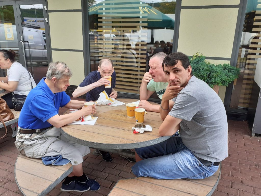 Czterech mężczyzn siedzących przy drewnianym stole na dworzu. Spożywają posiłek oraz piją kawę.