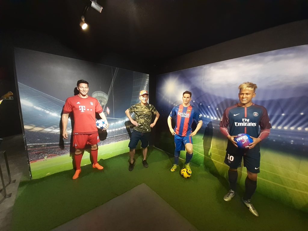 Ujęcie pokazuje trzy figury woskowe piłkarzy Roberta Lewandowskiego, Lionela Messi, Neymara oraz podopiecznego który z założonymi rekoma na biodra stoi między nimi.