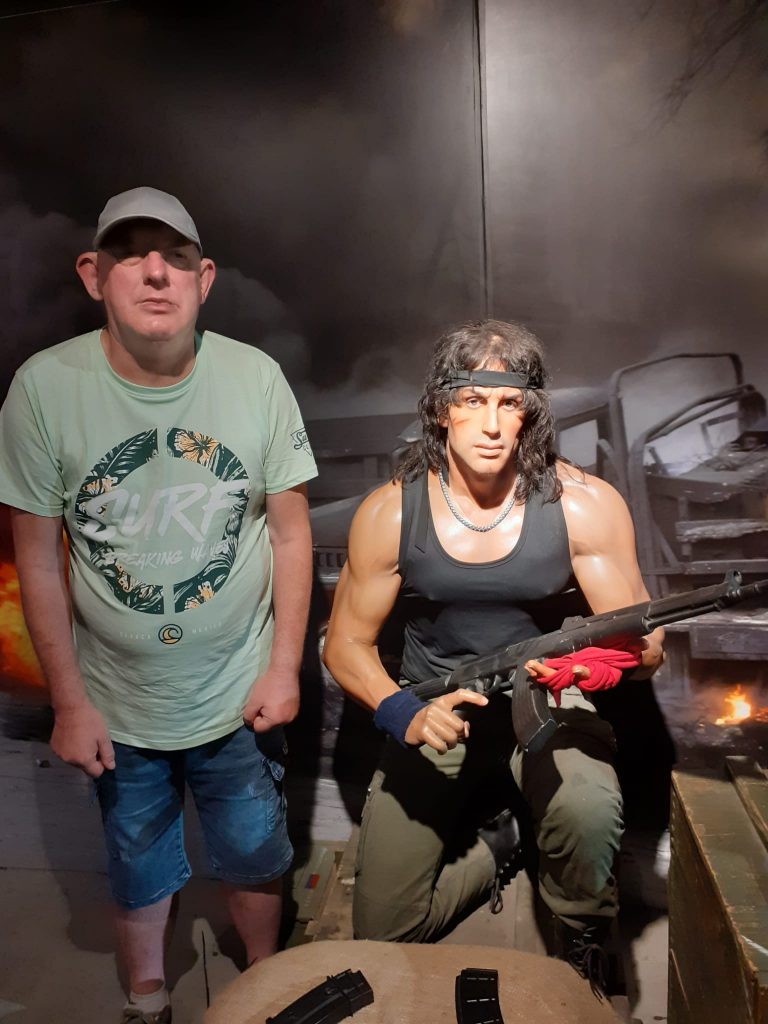 Podopieczny stoi obok postaci z firmu Rambo, która klęczy, ubrana jest w czarny podkoszulek, opaskę na głowie oraz karabin w rękach.