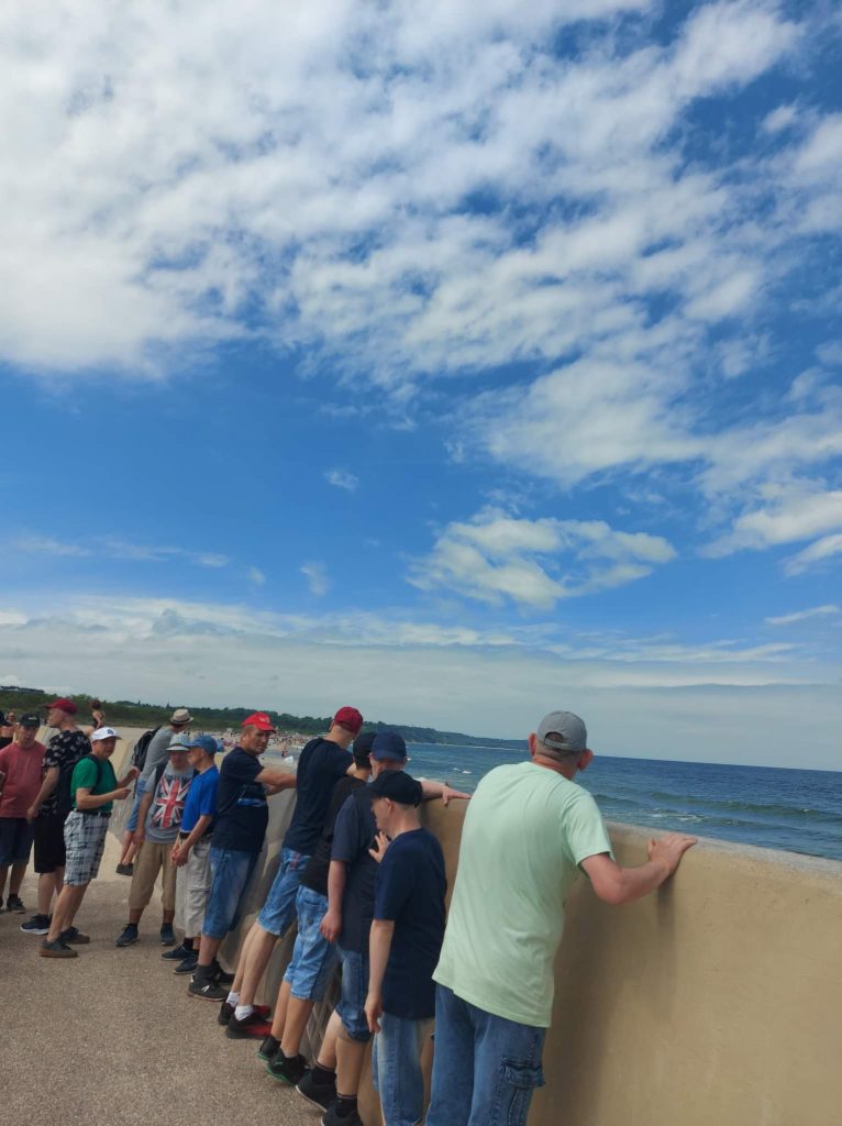 Grupa mieszkańców spoglądająca przez mur w stronę morza. Nad nimi niebieskie niebo z białymi chmurami.