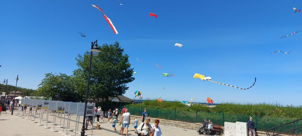 Deptak koło morza, po deptaku chodzą ludzie na niebie latają kolorowe latawce.