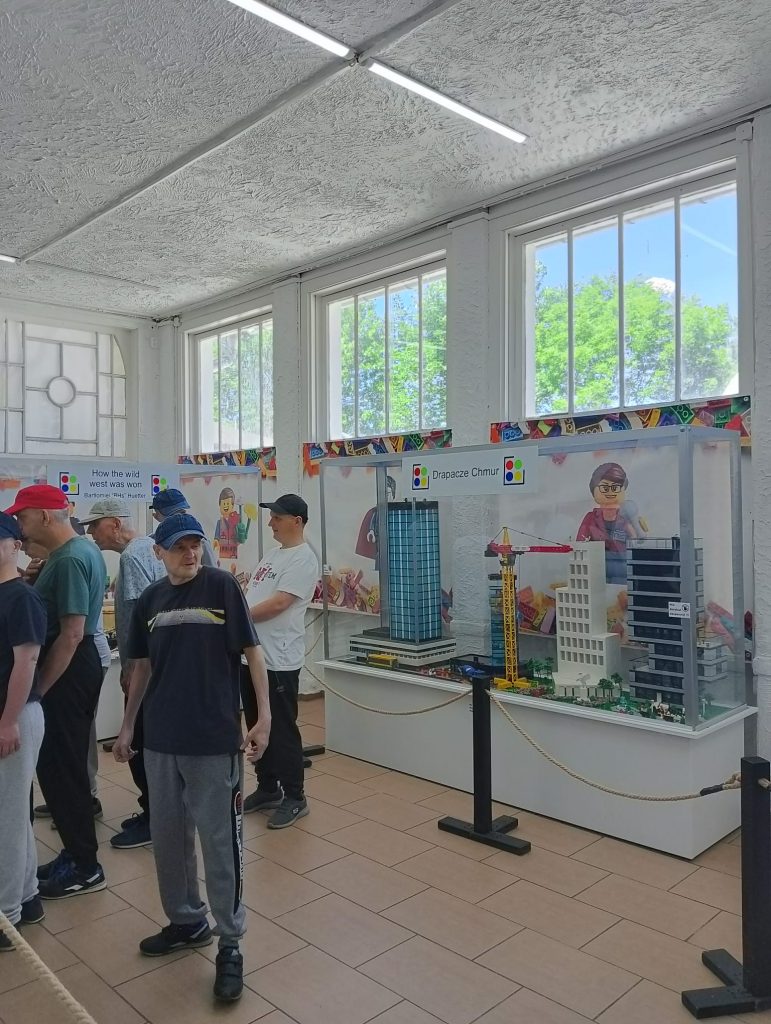Grupa mężczyzn stojąca w zamkniętymi pomieszczeniu, oglądają gabloty z wystawą klocków lego.
