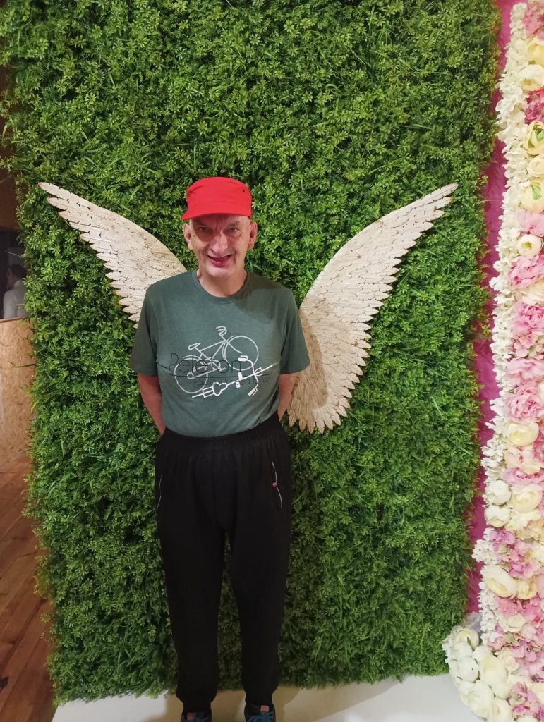 Mężczyzna ubrany w spodnie dresowe, koszulkę z nadrugiem oraz rowerem i czerwoną czapkę stoi na tle ściany z zielonymi roślinami na ścianie są skrzydła anioła.