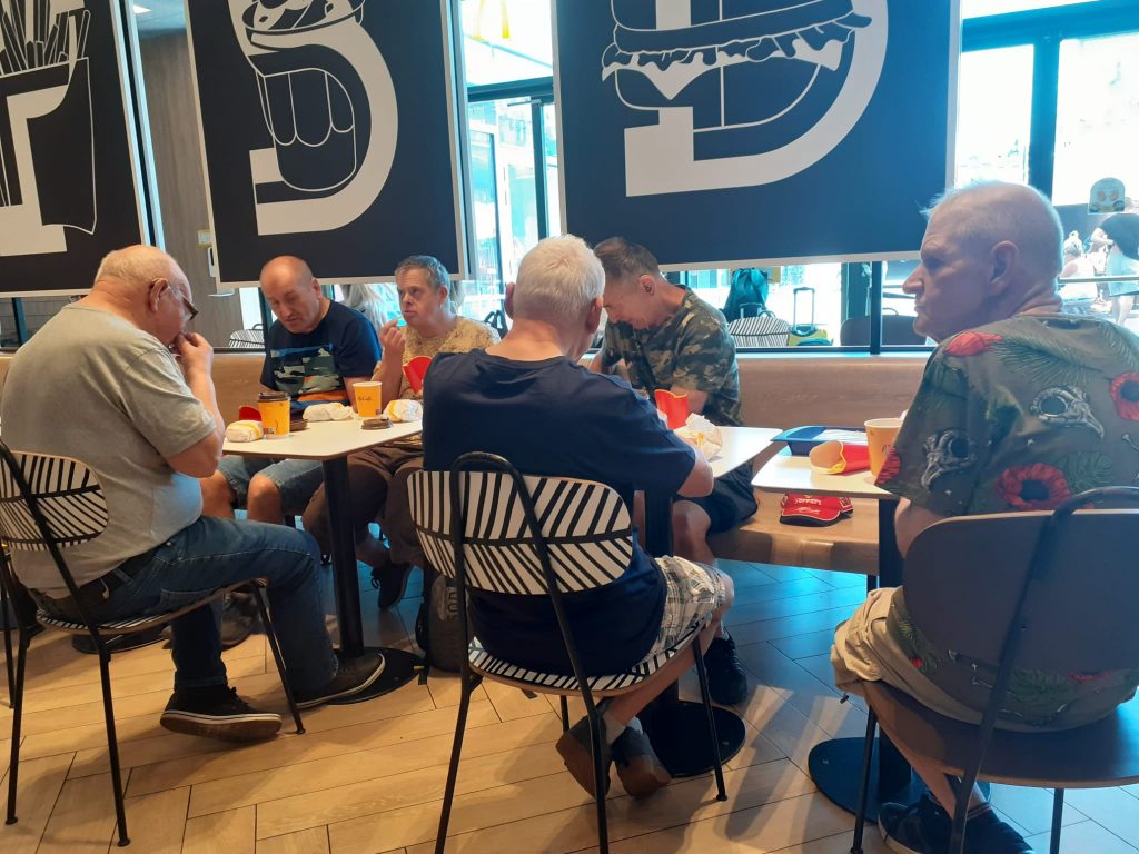 Sześciu podopiecznych siedzących przy stolikach wspólnie spożywających posiłek (frytki, burger oraz kawa).