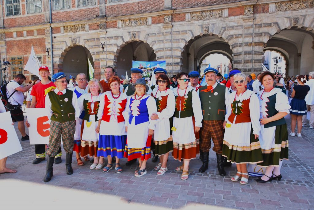 Grupa osób z kola gospodyń wiejskich ubrane w tradycyjne kolorowe Kociewskie stroje stoi przed bramą w Gdańsku.