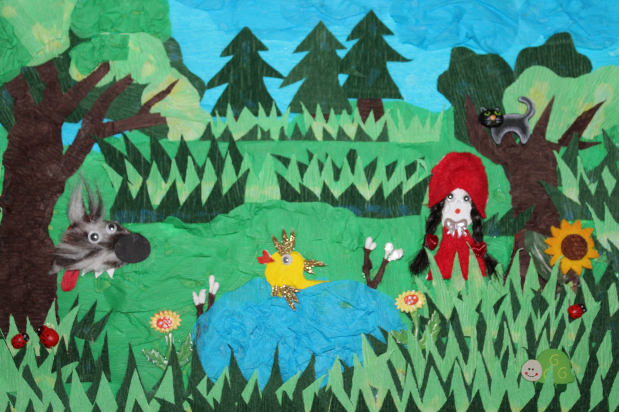 Obraz klejony z różnych elementów przedstawia wilka czerwonego kapturka, kota na drzewie oraz złotą rybkę w koronie. Wszyscy są w lesie, rybka pływa w stawie. Na trawce, czerwone biedronki.