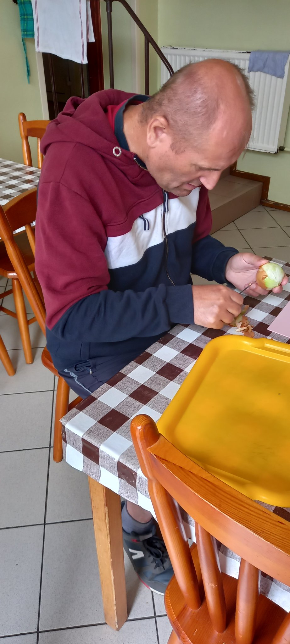 Mężczyzna ubran w bluzę dresową i krótkie sportowe spodenki siedzi przy stole gdzie obiera cebulę.