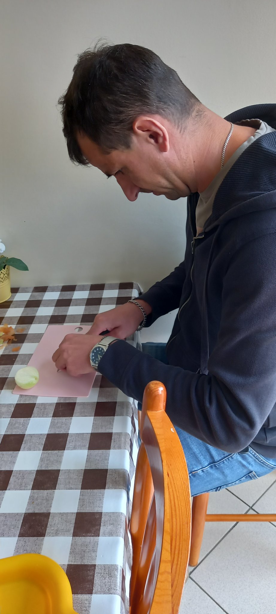 Mężczyzna siedzi w pomieszczeniu przy stole z ceratą w kratkę, kroi cebulę na desce.