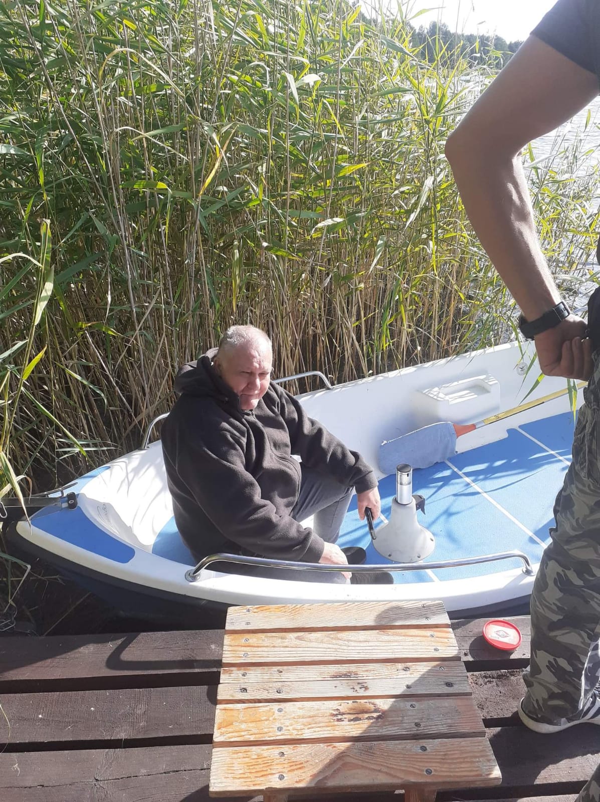 Mężczyzna siedzący w łódce w trzcinach przy pomoście. Nad nim widać postać mężczyzny w spodniach moro.