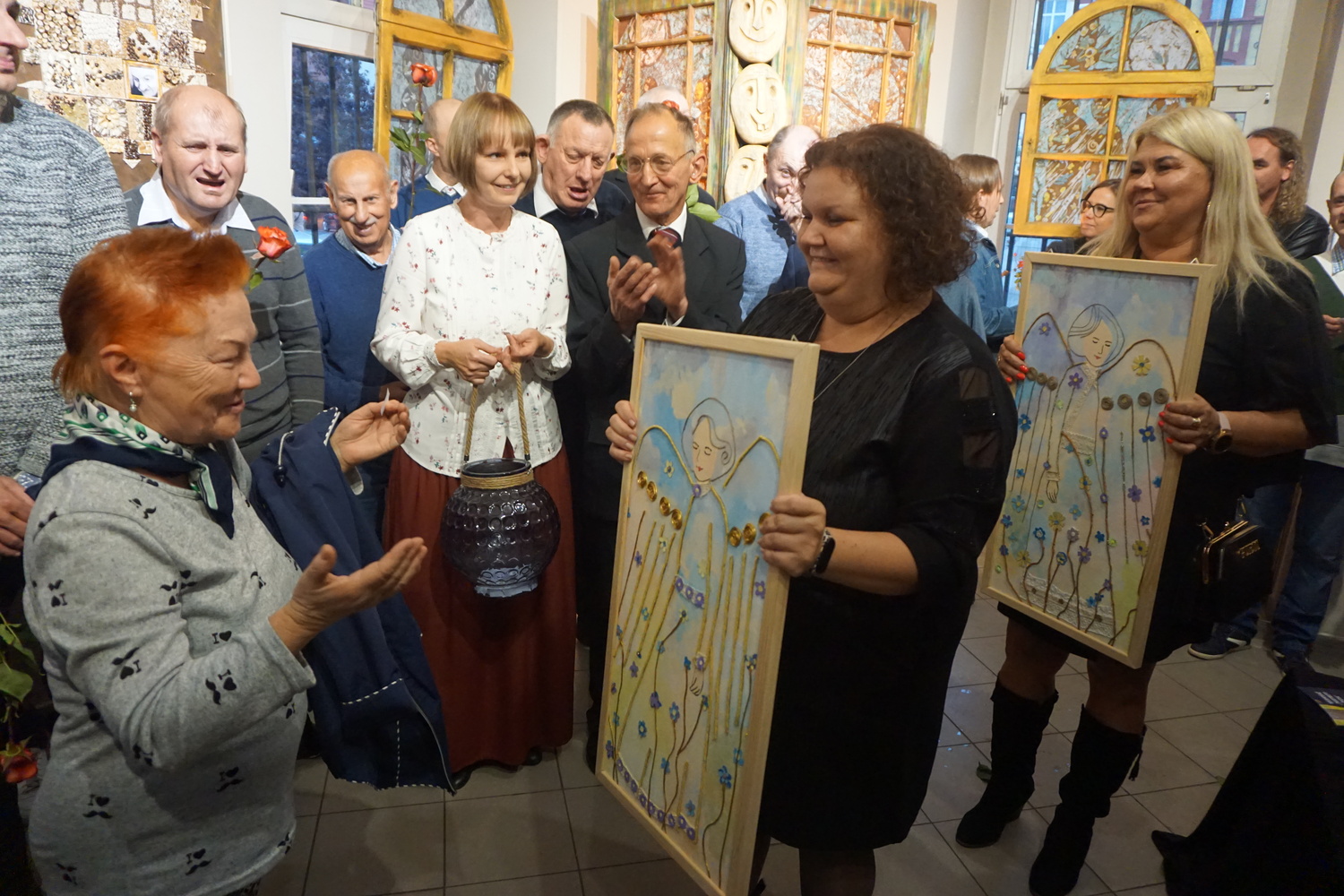 Kobieta stojąca na sali z rozłożonymi rękoma przed nią dwie inne osoby trzymające obrazy z aniołami. Jedna z nich wręcza obraz wygranej.