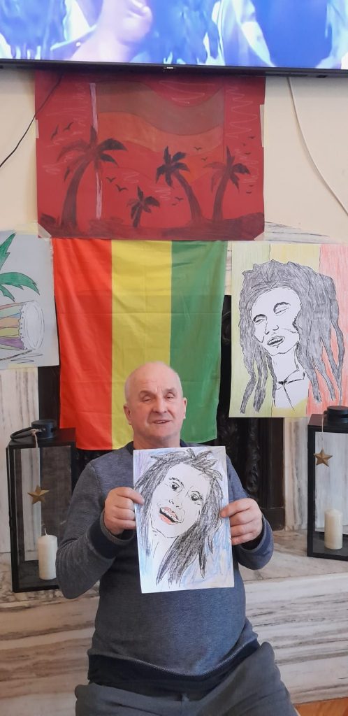 Uśmiechnięty podopieczny siedzi przed kominkiem i telewizorem. W tle palmy, obraz Boba Marleya i flaga. W ręce trzyma podobiznę artysty.