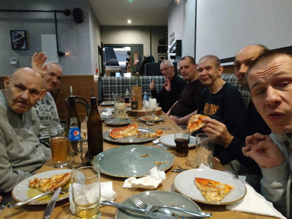 Uśmiechnięci mężczyźni siedzący przy stole, wspólnie jedzą posiłek - pizza. Do posiłku piją pepsi, soki oraz piwko. Spoglądają w stronę obiektywu.