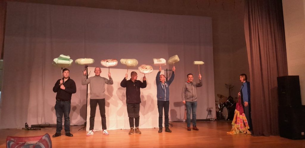 Pięciu mężczyzn podczas próby na scenie trzymają w rękach w górze artefakty teatralne. Przy zasłonie teatralnej stoi kobieta.