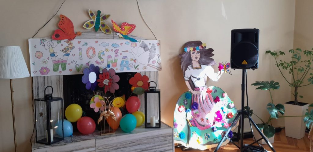 Kominek w pomieszczeniu z napisem Wiosna, pod nim kolorowe balony, kwiatki oraz motylek. obok stoi namalowana kobieta (marzanna) oraz głośnik na statywie
