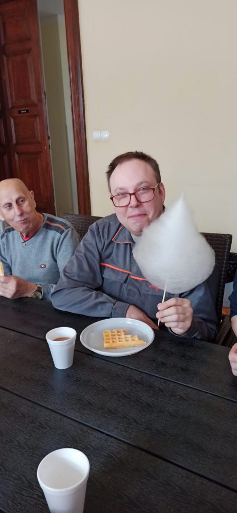 Mężczyzna siedzący przy stole trzyma w ręku watę cukrową. Przed nim leży talerzyk z gofrem oraz kawa. Obok inny mężczyzna spogląda na niego.