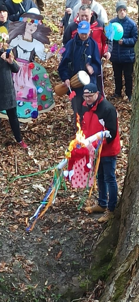 Instruktor terapii zajęciowej trzyma płonącą marzannę. W tle mężczyzna z bębenkiem oraz inne osoby.