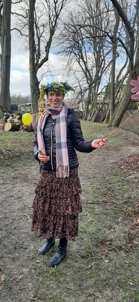 Uśmiechnięta kobieta z wiankiem na głowie spogląda w stronę obiektywu, w ręce trzyma sztuczny kwiat.