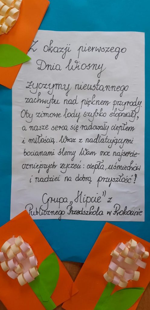 Grupa osób stojąca wspólnie w pracowni terapii zajęciowej. Na stole drewniane króliczki, farby oraz inne przedmioty. Dwie osoby trzymają prezent jedna z kobiet trzyma kwiaty. Wszyscy pozują do zdjęcia.