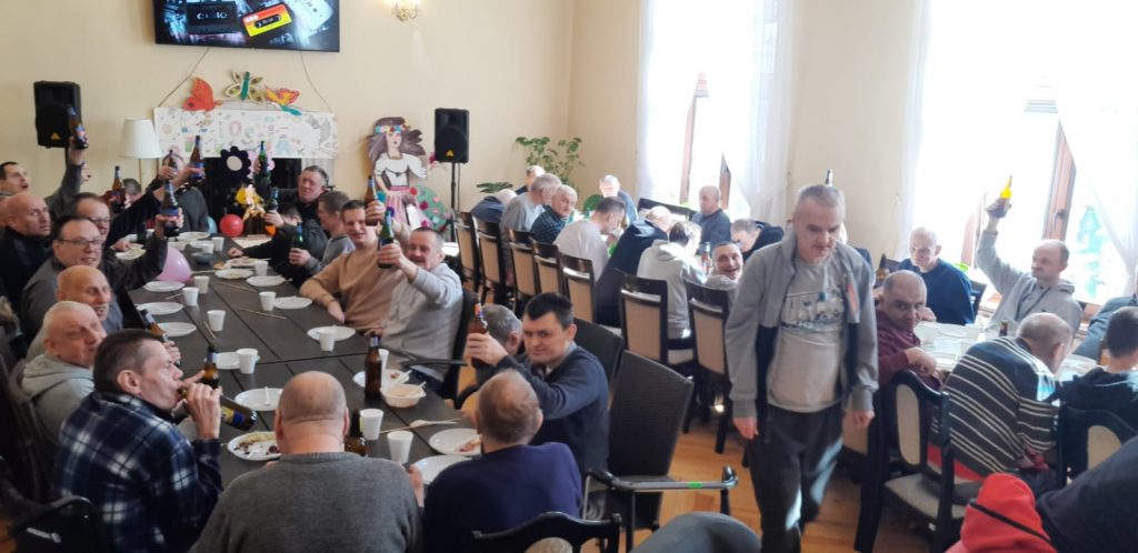 Grupa mężczyzn siedząca przy dwóch długich stołach trzymają w rękach butelki (bezalkoholowe piwo) na stole plastikowe talerzyki oraz kubeczki.