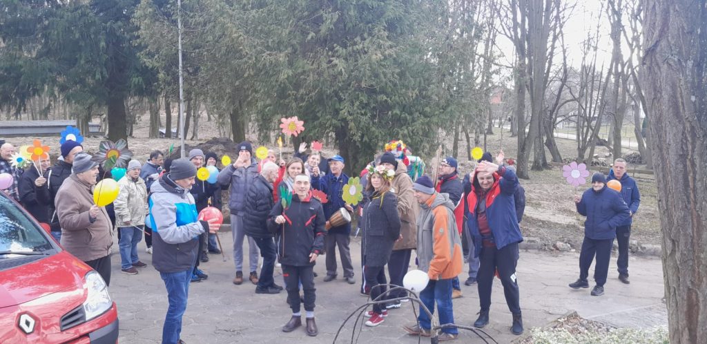 Grupa mężczyzn ubranych w kurtki, czapki trzymających kolorowe sztuczne kwiaty oraz balony.