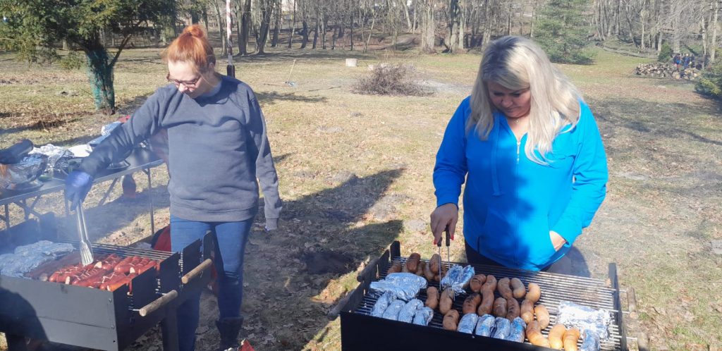 Dwie kobiety stojące przy dwóch grillach, przekładają kiełbaski. W tle park.