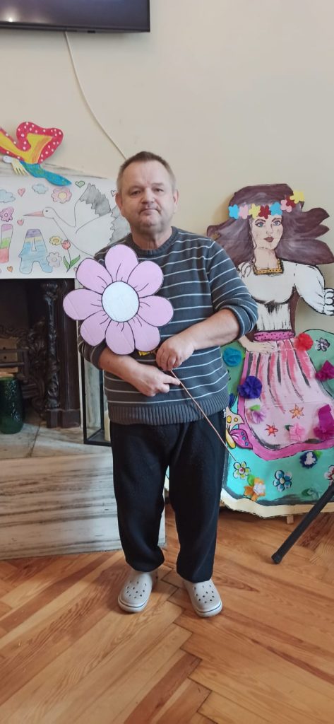 Mężczyzna stojący z sztucznym kwiatem w pomieszczeniu. Za nim ozdobiony kominek oraz sztuczna narysowana na kartonie kobieta (Marzanna).