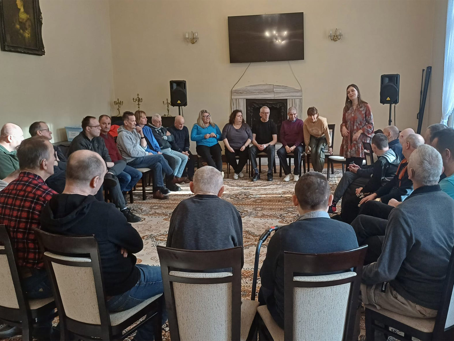 Grupa mieszkańców siedząca w kółku na krzesłach, w kole stoi kobieta.